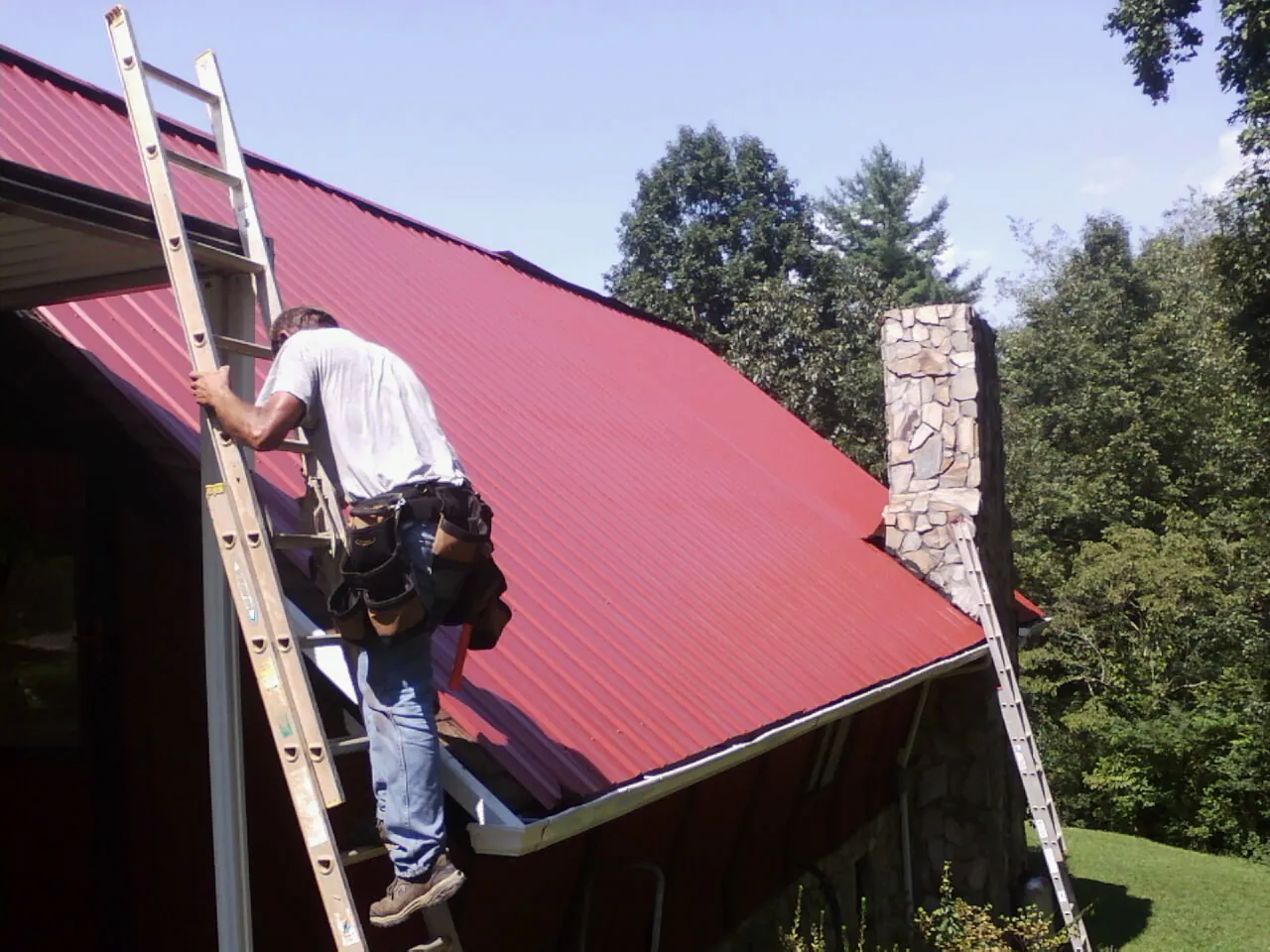 Roofer on a ladder beside a red metal roof and stone chimney — Soffit & Fascia Repair by Roofing Brothers in Putney